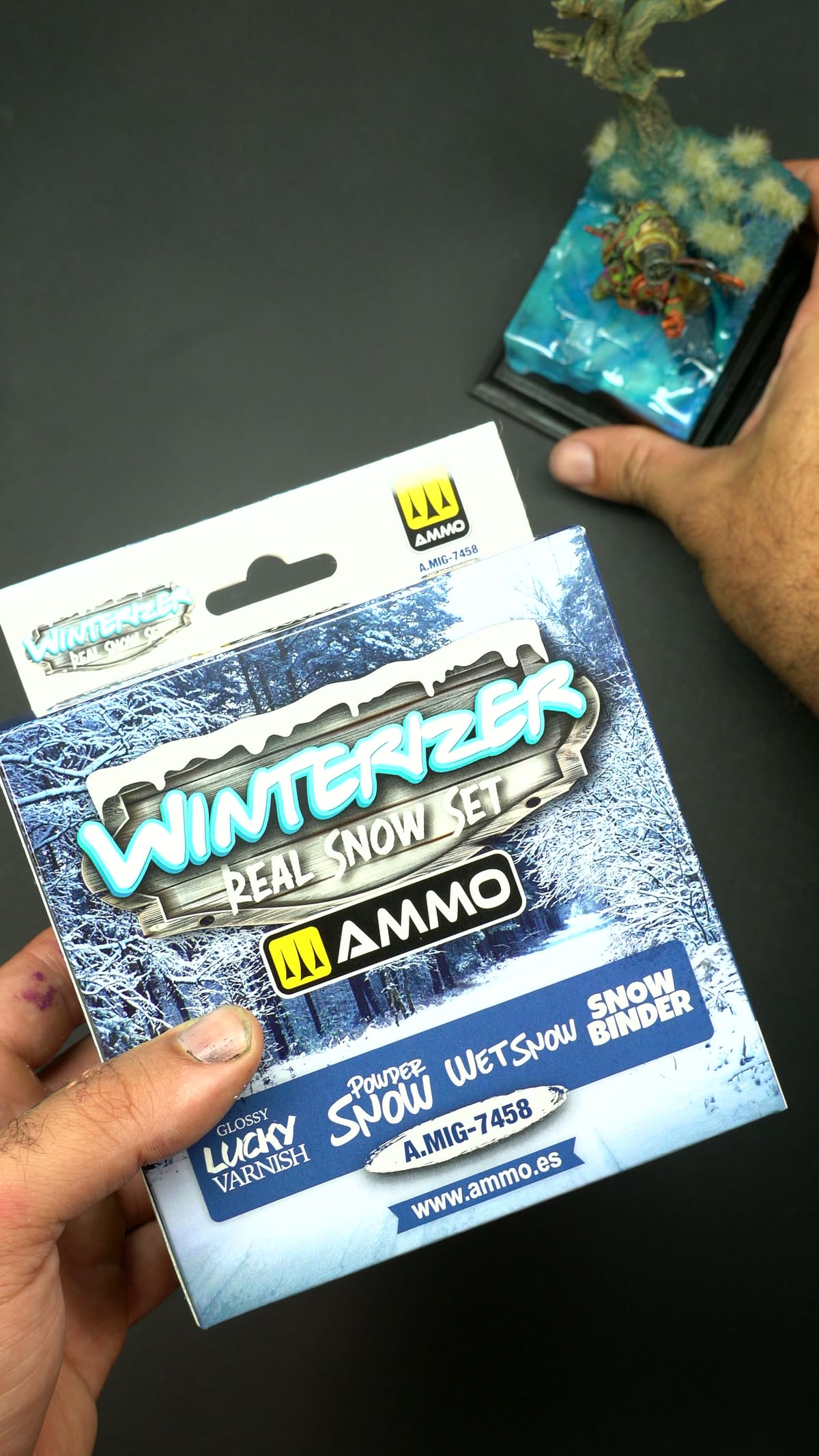 Une main tenant la boîte du Winterizer Real Snow Set d’AMMO, la figurine en arrière-plan