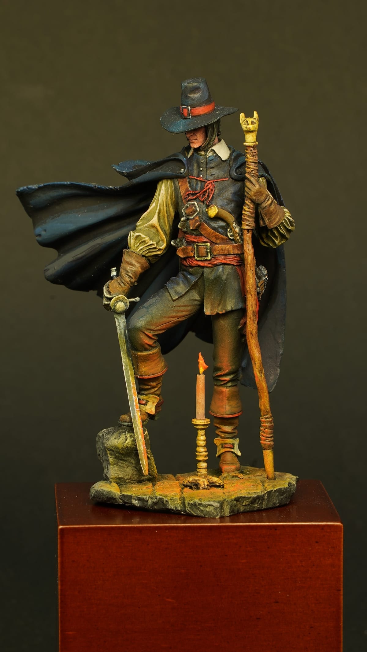 A figure in a wide-brimmed hat and long cloak, with a staff, on a red plinth