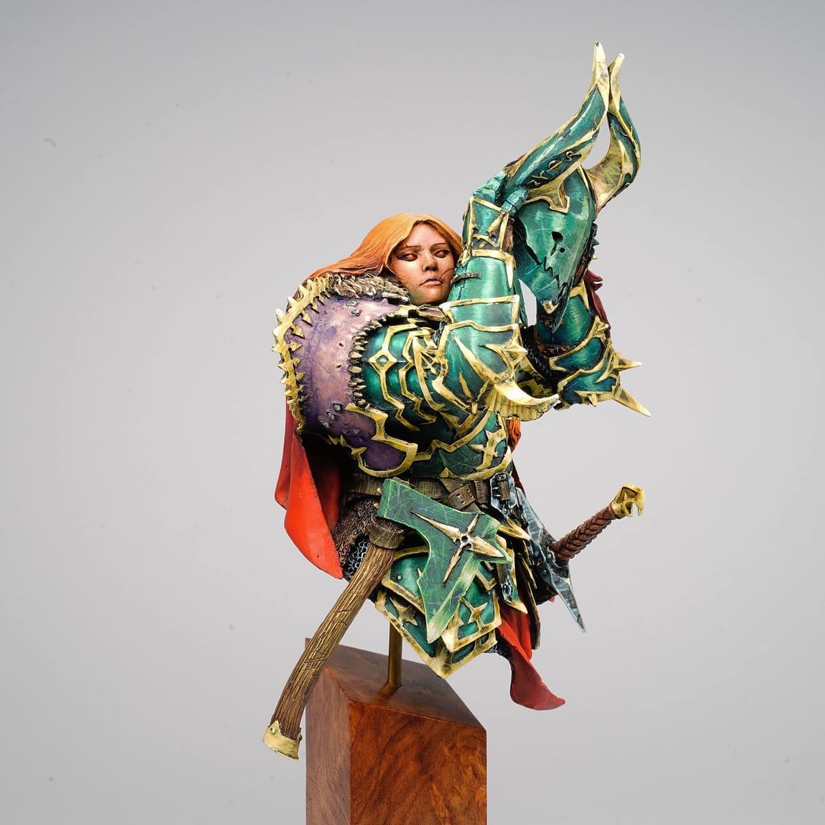Busto de Yrsa Grym: guerrera pelirroja con armadura turquesa y dorada, sobre un pedestal de madera